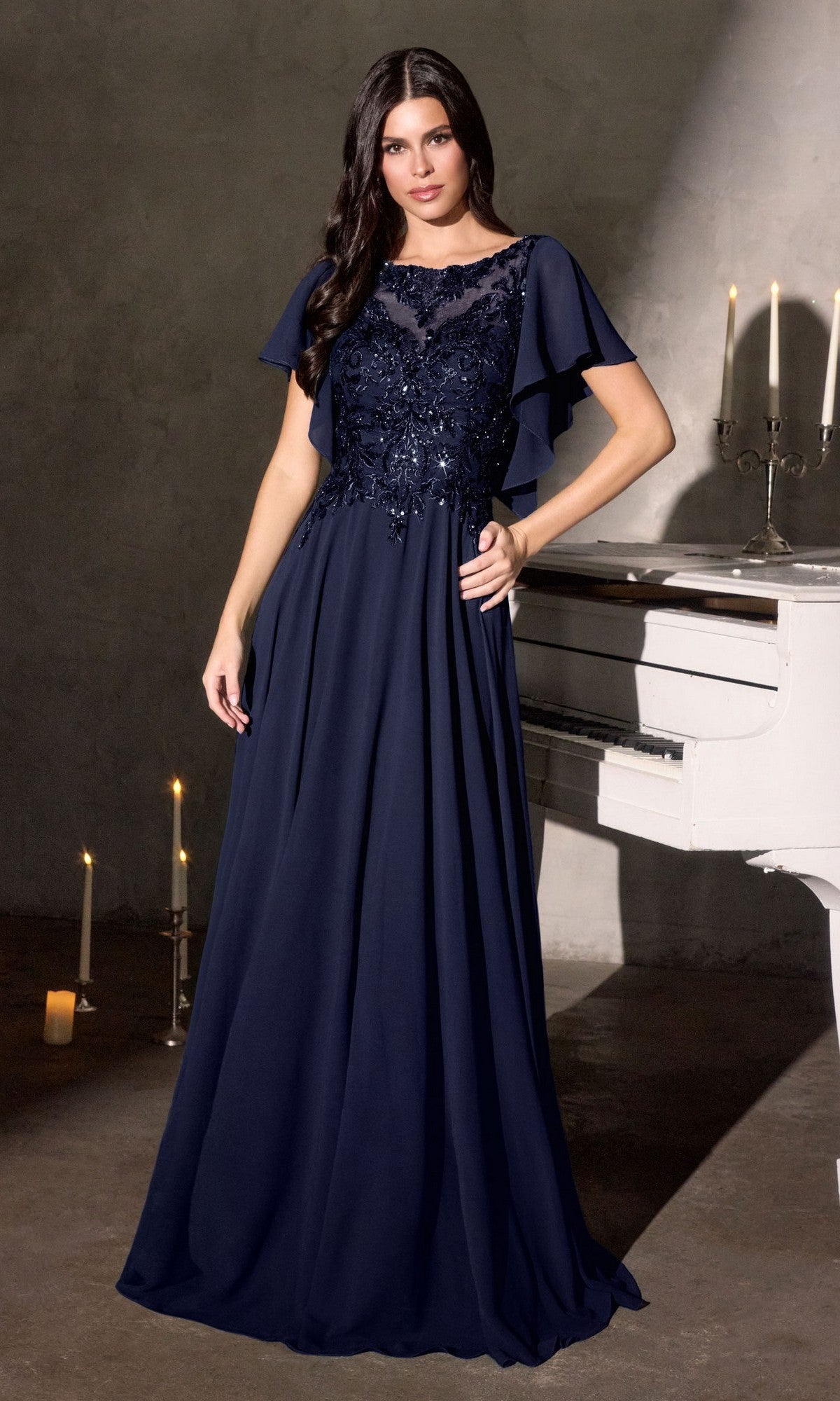 A woman in a navy Ladivine CL09 long formal dress stands by a white piano in a dimly lit room decorated with candles.